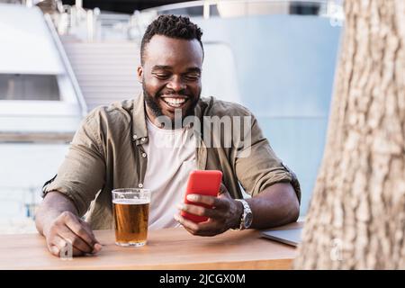 Glücklicher afrikanischer Mann, der Bier an der Brauerei-Bar trinkt, mit Mobiltelefon im Freien und einer Luxusyacht im Hintergrund - Fokus auf Gesicht Stockfoto