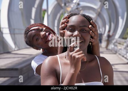 Zwei afroamerikanische Frauen bedeckten ihre Augen und umarmten sich von hinten. Zwei überraschte Mädchen. Frauen, die Spaß haben und Gesichtsemotionen zeigen Stockfoto