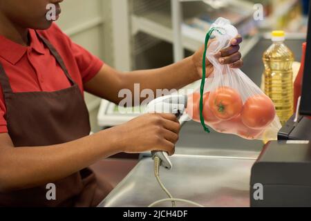 Junge schwarze Frau in rotem Hemd und brauner Schürze scannt frische Tomaten in Zellophanbeutel über den Kassenschalter im Supermarkt Stockfoto