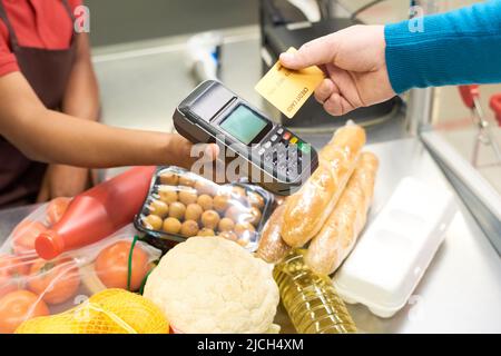 Männliche Verbraucher, die für Lebensmittelprodukte per Kreditkarte bezahlen, während sie sie über das Zahlungsterminal in der Hand der Verkäuferin am Schalter halten Stockfoto