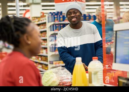 Glücklicher junger afroamerikanischer Käufer in Casualwear, der die Verkäuferin an der Theke sieht und Lebensmittel scannt Stockfoto