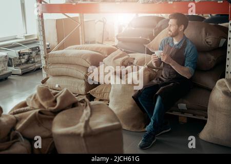 Geschäftsinhaber mit einer Tasse Kaffee und einem Telefon sitzen im Lager der Kaffeefabrik und schauen weg Stockfoto