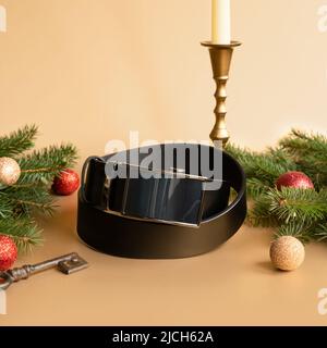 Aufgerollter schwarzer modischer Herren Ledergürtel mit automatischer Schnalle, Weihnachtstanne, glänzenden Kugeln, goldenem Kerzenständer und Schlüssel auf goldenem Hintergrund. Stockfoto