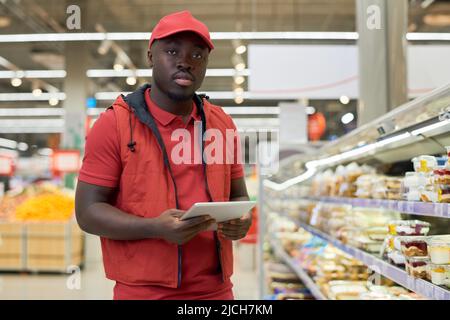 Ernsthafter junger schwarzer Mann mit Tablet, der vor einem großen Display steht und Desserts in Plastikbehältern im Supermarkt verpackt Stockfoto
