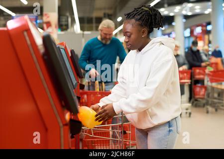 Junge Verbraucherinnen scannen eine Flasche Saft, während sie das Self-Service-Terminal gegen reife männliche Kunden im modernen Supermarkt nutzen Stockfoto