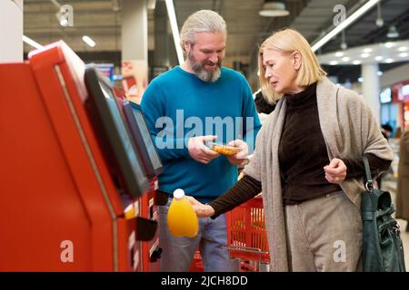 Reife blonde Verbraucherin scannt Flasche Orangensaft im Terminal der Selbstbedienung, während sie Lebensmittel im Supermarkt kauft Stockfoto