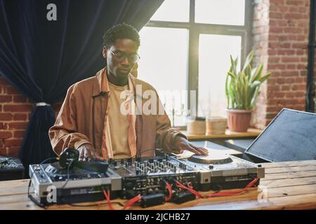 Junger ernsthafter schwarzer Mann, der vor der Aufführung auf das dj-Board schaut, während er musikalische Ausrüstung adjustisch anfasst und Plattenspieler berührt Stockfoto