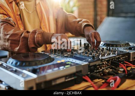 Hände von modernen schwarzen Mann, die Klänge auf dj-Set regulieren, während sie am Tisch im Studio oder Loft-Apartment stehen und Mischer drehen Stockfoto