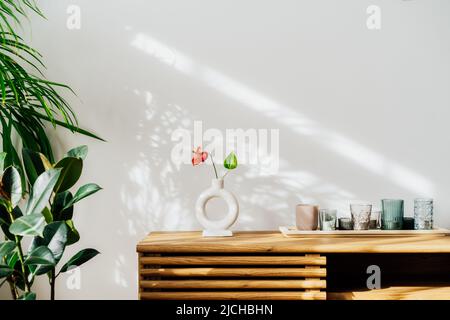 Moderne Einrichtung im minimalistischen skandinavischen Stil. Nordische Keramikvase mit roter Anthuriumblume, Kerzen auf einer Holzkonsole. Grünhaus Pflanzen in Töpfen Stockfoto