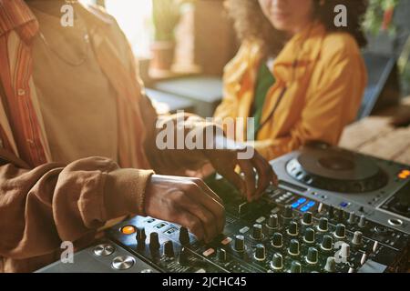 Hände von afroamerikanischem Kerl im Sweatshirt, der DJ-Controller anpasst, während er neue Musik für ein Unterhaltungsevent gegen seine Freundin kreiert Stockfoto