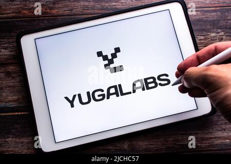 Das Logo von Yuga Labs (der Muttergesellschaft des Bored Ape Yacht Club) auf einem Tablet auf einem Holztisch. Handgriff mit kabellosem Stylus Stockfoto