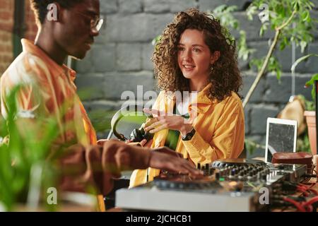 Lächelndes Mädchen mit Kopfhörern in den Händen, das ihren Freund beim Mischen von Geräuschen ansieht, während er mit dem dj-Controller im Studio am Tisch steht Stockfoto