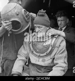 1950s, historisch, ein Tiefseetaucher in der Ausrüstung des Tages, sitzend, mit seinem schweren Messingtauchhelm auf den Kopf gesetzt, im Hafen von London, England, Großbritannien. Stockfoto