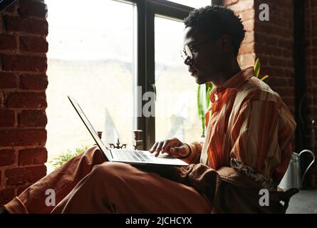 Seitenansicht eines jungen schwarzen Mannes, der auf den Laptop-Bildschirm schaute, während er in einer Loft-Wohnung am Fenster saß und sich mit ihm vernetzte Stockfoto