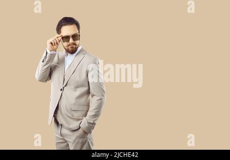 Geschäftsmann in elegantem Anzug und Sonnenbrille posiert auf leerem beigefarbenem Kopierraum-Hintergrund Stockfoto