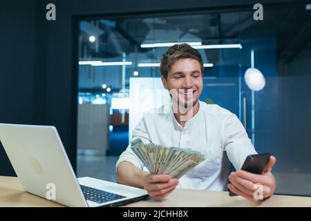Der Mann arbeitet im Büro, der Geschäftsmann hält Dollar Bargeld freut sich und lächelt schaut auf den Telefonbildschirm, freut sich über den Sieg Stockfoto