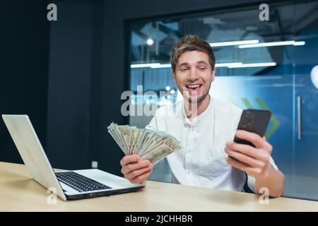 Der Mann arbeitet im Büro, der Geschäftsmann hält Dollar Bargeld freut sich und lächelt schaut auf den Telefonbildschirm, freut sich über den Sieg Stockfoto