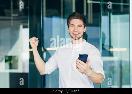 Porträt eines erfolgreichen Geschäftsmannes Mann schaut auf die Kamera und lächelt, hält ein Telefon, freut sich über Erfolg und Triumph, feiert Geste Stockfoto