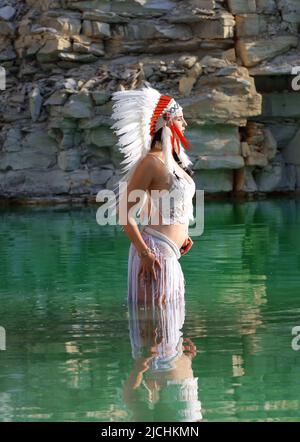 Ein Mädchen wird als Indianerin der Ureinwohner angesehen. Sie ist in Weiß gekleidet und trägt einen gefiederten Kopfschmuck. Man sieht sie in einem Steinbruch-See stehen. Stockfoto