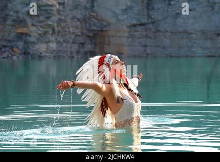 Ein Mädchen wird als Indianerin der Ureinwohner angesehen. Sie ist in Weiß gekleidet und trägt einen gefiederten Kopfschmuck. Man sieht sie in einem Steinbruch-See stehen. Stockfoto