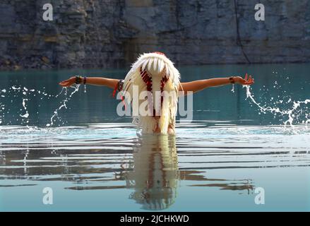 Ein Mädchen wird als Indianerin der Ureinwohner angesehen. Sie ist in Weiß gekleidet und trägt einen gefiederten Kopfschmuck. Man sieht sie in einem Steinbruch-See stehen. Stockfoto