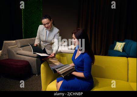 Eine hispanische Frau, angenehme Einzelhandelsmanagerin in einem Möbelgeschäft, die einen neuen Polsterkatalog vorstellt und Farbstoffmuster einem Kunden zeigt, der ich bin Stockfoto