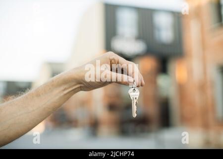 Hausschlüssel in männlicher Hand auf dem Hintergrund von Neubauten und Himmel. Immobilienmakler, Umzug oder Vermietung von Immobilien. Die Schlüsselübergabe einer Wohnung wird von einem Mann vor dem Hintergrund von Gebäuden gehalten. Stockfoto