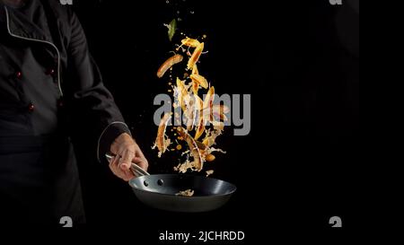 Anonymer Koch in Uniform wirft appetitlich gebratenes Schupfnudeln mit Würstchen in der Bratpfanne vor schwarzem Hintergrund während des Kochvorgangs im Studio Stockfoto