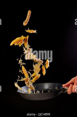 Crop anonyme Koch Spiegeln appetitlich gebratene Knödel und Würstchen in Metall-Bratpfanne während Kochen vor schwarzem Hintergrund im Studio Stockfoto