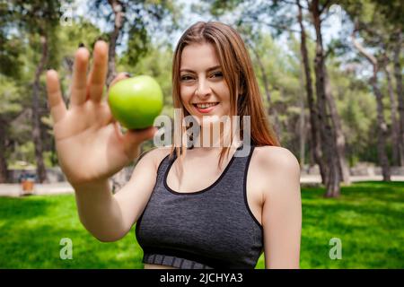 Fröhliche sportliche Frau im Sport-BH, die auf dem Stadtpark steht, lächelt im Freien, hält einen Apfel und blickt auf die Kamera. Outdoor-Sport und Gesundheit Stockfoto