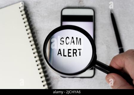 Die Hand eines Mannes hält eine schwarze Lupe am Griff auf einem Smartphone-Hintergrund mit einem Betrugswarntext. Stockfoto
