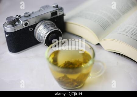Gebrauter grüner Kräutertee in einer transparenten Tasse neben einem offenen Buch und einer Vintage-Kamera auf hellem Hintergrund Stockfoto