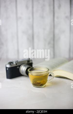 Gebrauter grüner Kräutertee in einer transparenten Tasse neben einem offenen Buch und einer Vintage-Kamera auf hellem Hintergrund Stockfoto