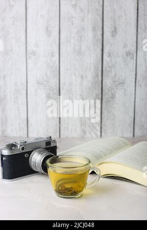 Gebrauter grüner Kräutertee in einer transparenten Tasse neben einem offenen Buch und einer Vintage-Kamera auf hellem Hintergrund Stockfoto