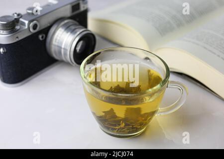 Gebrauter grüner Kräutertee in einer transparenten Tasse neben einem offenen Buch und einer Vintage-Kamera auf hellem Hintergrund Stockfoto