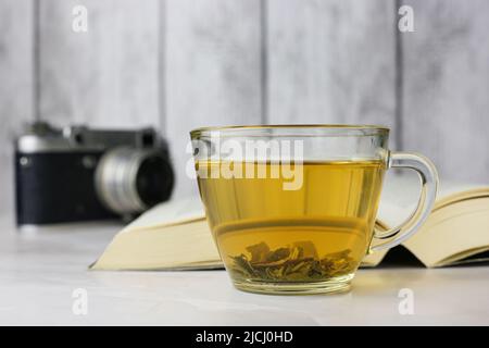 Gebrauter grüner Kräutertee in einer transparenten Tasse neben einem offenen Buch und einer Vintage-Kamera auf hellem Hintergrund Stockfoto