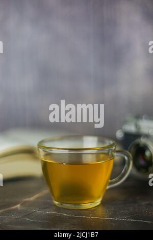 Gebrauter grüner Kräutertee in transparenter Tasse in der Nähe von offenem Buch und Vintage-Kamera auf dunklem Hintergrund Stockfoto