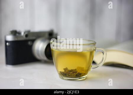 Gebrauter grüner Kräutertee in einer transparenten Tasse neben einem offenen Buch und einer Vintage-Kamera auf hellem Hintergrund Stockfoto