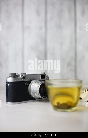 Gebrauter grüner Kräutertee in einer transparenten Tasse neben einem offenen Buch und einer Vintage-Kamera auf hellem Hintergrund Stockfoto