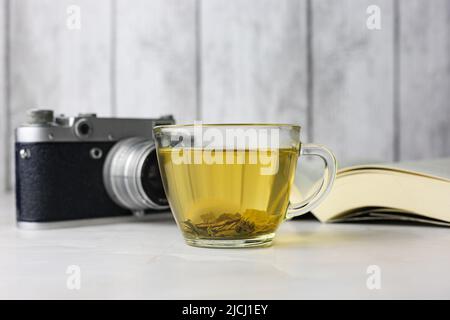 Gebrauter grüner Kräutertee in einer transparenten Tasse neben einem offenen Buch und einer Vintage-Kamera auf hellem Hintergrund Stockfoto