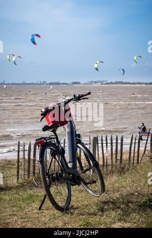 Schwarzes Elektro-Fahrrad mit einem roten Korb im Gras gegen Meer geparkt. Viele Kitesurfer reiten auf dem Meer unter einem klaren blauen Himmel Stockfoto