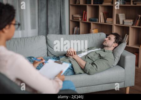 Unglücklicher trauriger europäischer Patient liegt auf dem Sofa, hört der schwarzen Psychologin im Inneren der Büroklinik zu Stockfoto