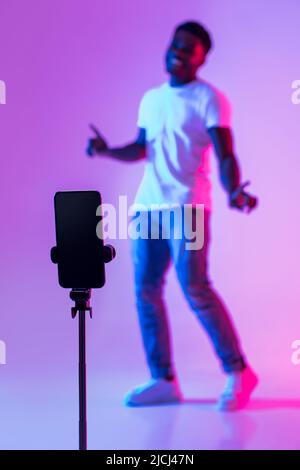 Schwarzer männlicher Vlogger, der Video für soziale Medien auf dem Handy schießt, Spaß im Neonlicht hat, Mockup Stockfoto