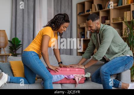 Vorbereitung auf den Urlaub. Aufgeregt schwarzes Paar packen und versuchen, Koffer auf dem Sofa zu schließen, Vorbereitung für den Sommerurlaub Stockfoto