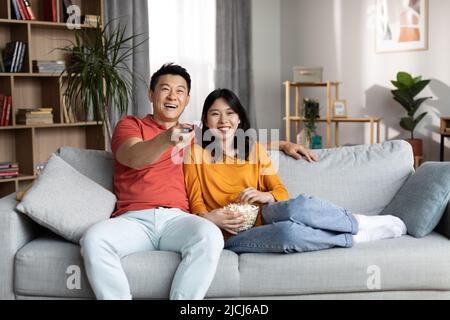 Liebendes chinesisches Paar, das sich auf dem Sofa ausruht, Fernsehen schaut und lacht Stockfoto