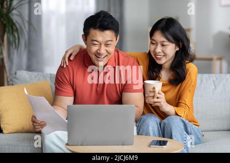 Lächelnde asiatische Ehegatten zahlen Rechnungen online, mit Laptop Stockfoto