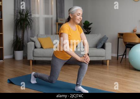 Positive sportliche Senior-Frau, die sich zu Hause fit hält, Ausfallschritte macht, im Wohnzimmer auf einer Yogamatte trainiert und lächelt Stockfoto