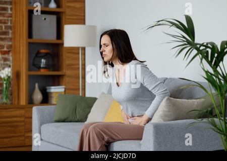 Kranke junge Frau, die an Menstruationsschmerzen leidet. Frau mit den Händen, die den Bauch quetschen, mit schmerzhaften Bauchschmerzen oder Periodenkrämpfen, die auf dem Sofa sitzen Stockfoto