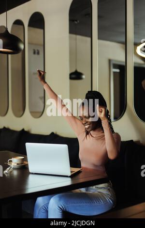 Froh erfolgreiche und stolze interracial afro amerikanische Geschäftsfrau auf Remote-Arbeit im Café. Den Arm heben, den Sieg Stockfoto