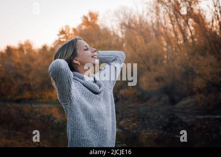 Nahaufnahme einer jungen blonden kaukasischen Frau, die im Wald steht und mit beiden Händen hinter dem Kopf lächelnd nach oben blickt, mit Bäumen, die mit Sonnenuntergang bedeckt sind Stockfoto